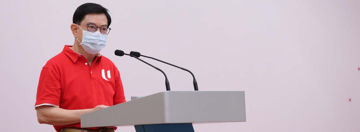 Lawrence Wong at podium, wearing mask and red polo shirt with a 'U' logo. Two microphones are present.